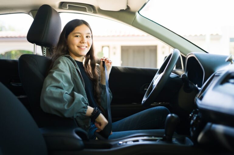 A teenage girl in a green jacket smiles as she sits behind the wheel of a car and buckles her seat belt.