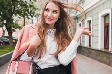 A woman in a white dress shirt, pink blazer, and black slacks smiles playfully and flicks her hair as she strolls down a sidewalk.
