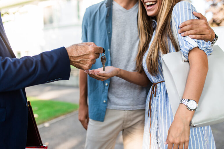 A person in a suit hands house keys to a smiling woman standing beside a man who has his arm around her shoulders.