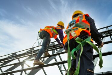 Two construction workers wearing orange safety vests and yellow hard hats climb up a steel enclosure.