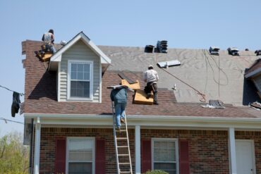 A group of workers on the roof of a home replacing the shingles using power tools with pneumatic hoses.