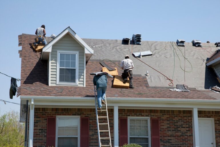 A group of workers on the roof of a home replacing the shingles using power tools with pneumatic hoses.