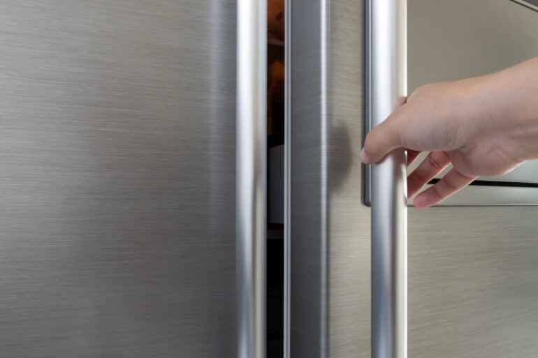 An extremely close-up view shows a single hand pulling the handle of a stainless steel refrigerator.