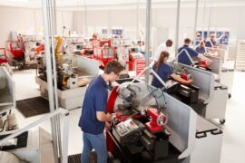 Six team members in blue shirts work at standing workstations in a factory. A person in a white shirt inspects their work.