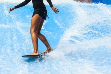 A person wearing a black long-sleeve bathing suit and black shorts stands on a board in a surf pool.