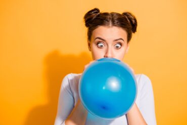 A woman with buns in her hair stands against an orange background, blowing up a blue balloon. She is looking at the balloon.