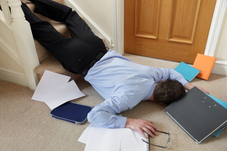 A man lies at the bottom of a stairway, having fallen. Folders and papers are scattered on the floor.