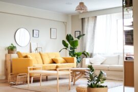 A home's living room contains a yellow couch, a white sofa, a tan rug, a wooden coffee table, and potted plants.