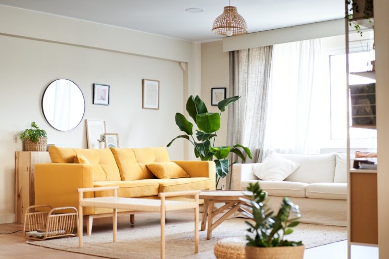 A home's living room contains a yellow couch, a white sofa, a tan rug, a wooden coffee table, and potted plants.
