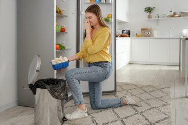 A woman kneels next to a garbage can and holds her nose as she throws away a bowl of eggs.