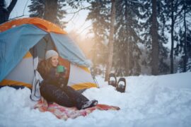 A woman sits in the opening of her tent and drinks from a mug. The tent is set up in a snowy forest.
