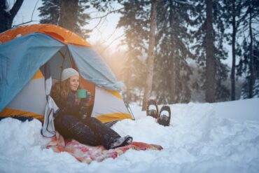 A woman sits in the opening of her tent and drinks from a mug. The tent is set up in a snowy forest.