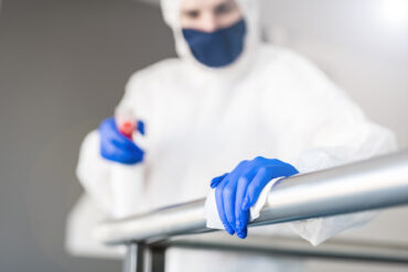 A professional cleaner in a white body armor suit and a face mask. They're wearing blue latex gloves to disinfect a railing.