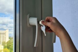 Close-up of someone inserting a key into a window to lock it. An apartment building is visible outside the window in the distance.