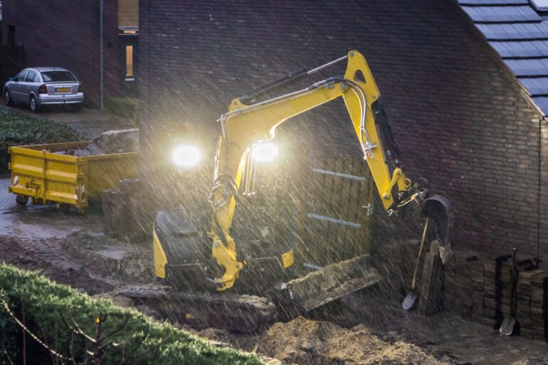A small backhoe digs up a plot of soil in heavy rain. Bright lights shine behind the backhoe.
