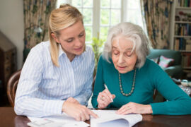 An elderly woman prepares to sign paperwork in pen, while a younger woman points to a section of the document.