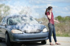 A woman stands and talks on her cell phone next to a blue car with smoke coming out from under the hood.