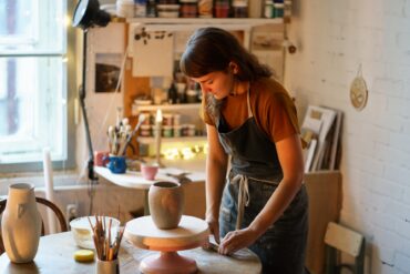 A woman stands next to a table, wearing a smock, and works with some clay. She's in an art studio around other ceramics.