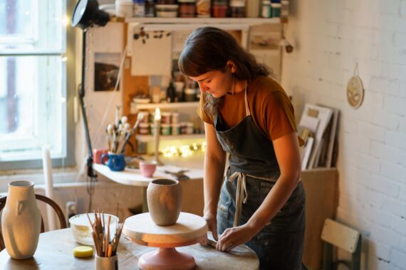 A woman stands next to a table, wearing a smock, and works with some clay. She's in an art studio around other ceramics.