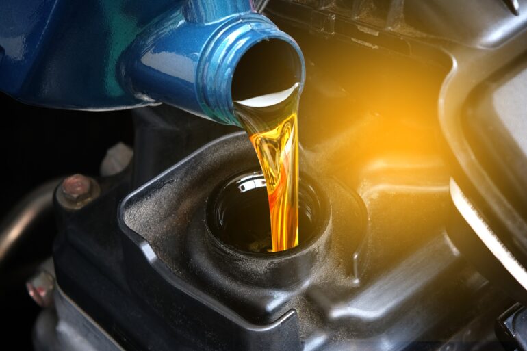 A close-up view shows a thick, yellow liquid from a blue bottle being poured into an opening on an engine.
