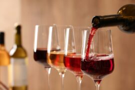 A dark-colored wine bottle pours a red liquid into a wine glass. Three other wine glasses full of wine sit next to it.