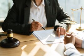 Two people sit across from each other at a desk. One points at a piece of paper while the other rests their hands.