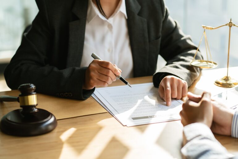 Two people sit across from each other at a desk. One points at a piece of paper while the other rests their hands.