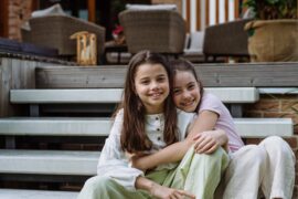 Two young girls, each with brown hair, sitting on the steps in front of a house. The two girls share an embrace.