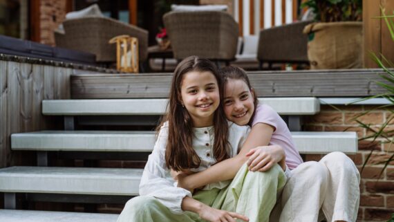 Two young girls, each with brown hair, sitting on the steps in front of a house. The two girls share an embrace.