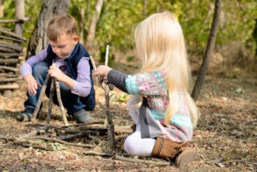 Two young children sitting on the ground outside in a wooded area, playing with sticks on a sunny day.