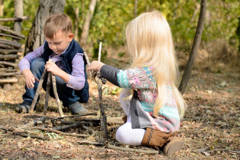 Two young children sitting on the ground outside in a wooded area, playing with sticks on a sunny day.