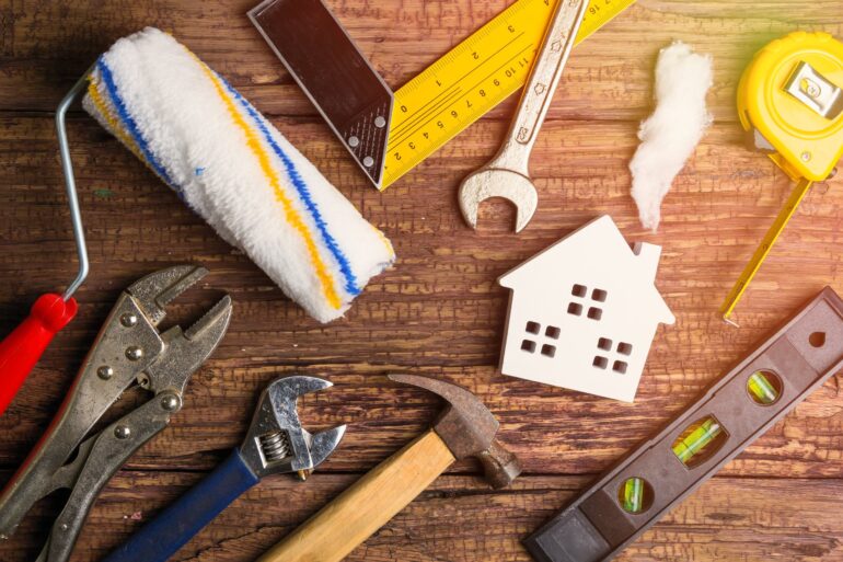 A hammer, paint roller, wrench, and other tools lay on a wooden table with a small white model of a home.