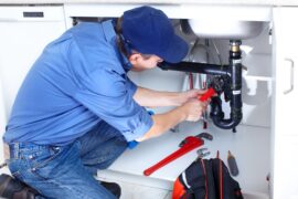 A plumber is on their knees and working on the pipes under a sink using a red wrench with other tools nearby.