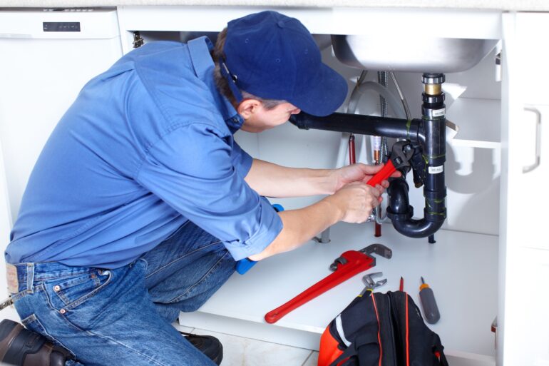 A plumber is on their knees and working on the pipes under a sink using a red wrench with other tools nearby.
