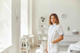 A woman wearing white scrubs stands with one hand in her pocket in a professional medical office.
