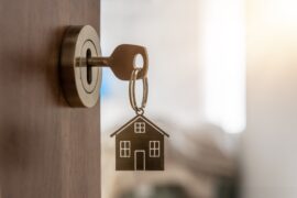 A key hangs out of a lock, the door slightly open. On the key is a keychain in the shape of a house.