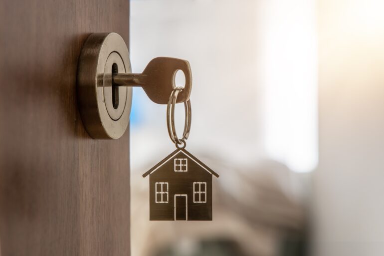A key hangs out of a lock, the door slightly open. On the key is a keychain in the shape of a house.