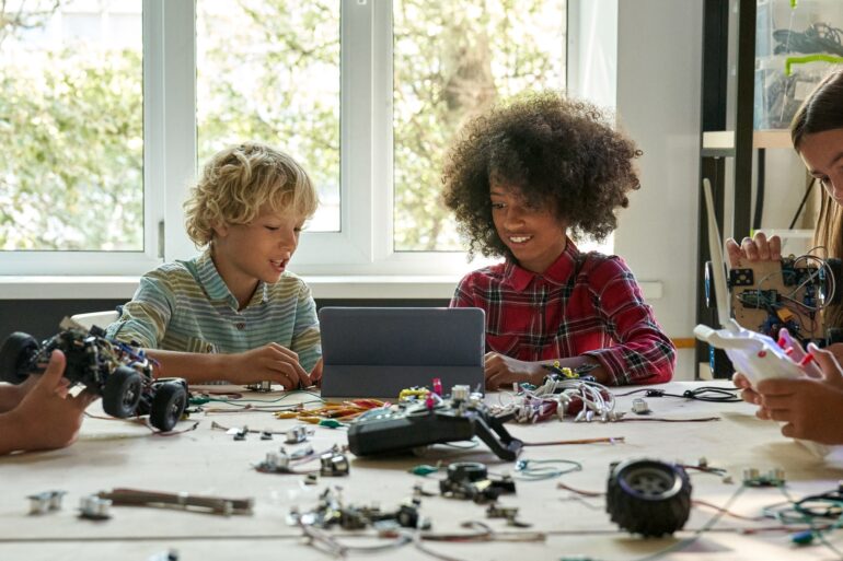 A group of pre-teen students gathered around a desk that is covered in the pieces of a hand-built robotic devices.