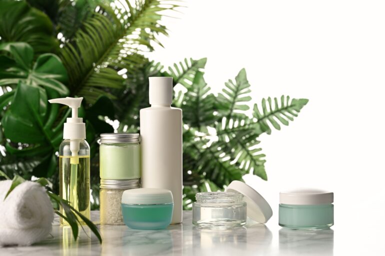 An arrangement of skin-care products laid out on a marble surface in front of a green, leafy fern plant.