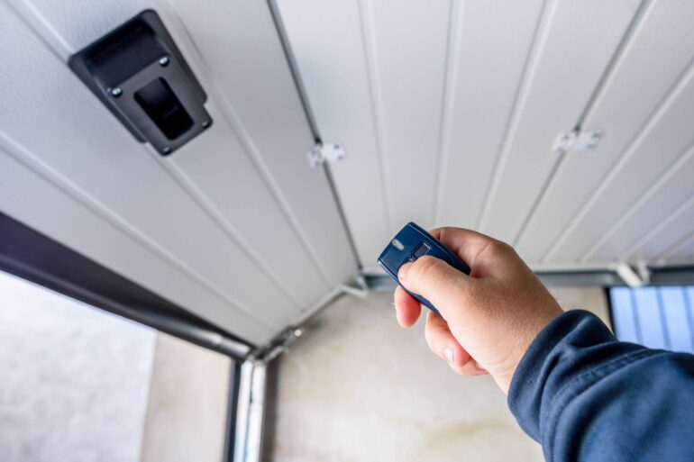 A person holds a navy blue remote with two grey buttons, pressing one while facing a white panel garage door.