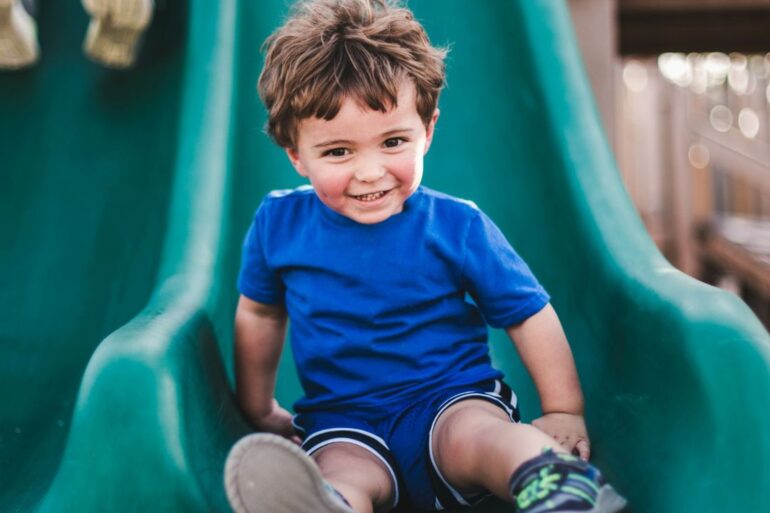 A toddler in a blue shirt sitting at the end of a plastic green slide in an outdoor playground space.