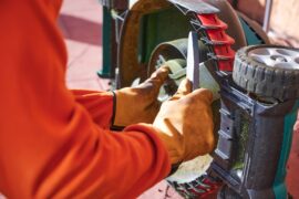 A close-up of a person wearing gardening gloves while sharpening the blades of a lawnmower with a grindstone.
