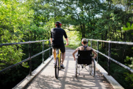 Two people, one on a bicycle and the other in a wheelchair, crossing a bridge surrounded by green trees in a park.
