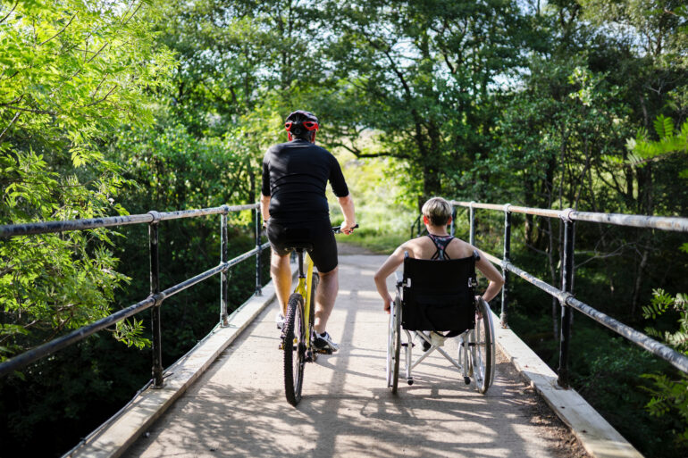 Two people, one on a bicycle and the other in a wheelchair, crossing a bridge surrounded by green trees in a park.