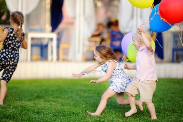 Three children running outside in a decorated backyard. They each hold colorful balloons in their hands.