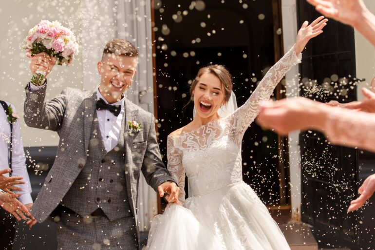 A smiling bride and groom standing side-by-side in a shower of rice and grains thrown by their guests.