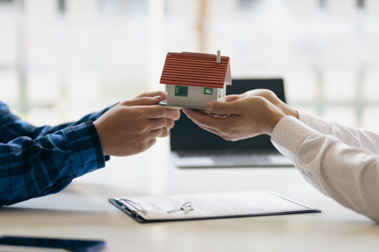 A close-up of a miniature home being transferred between two pairs of hands across a table above a contract.