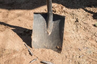 A close-up view shows a dark-colored shovel stuck in sandy soil on a bright, sunny day. A stick appears near the shovel.