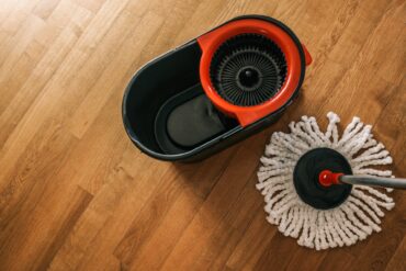 A spin mop system on the wood flooring. The mop head is sherpa with a black and red bucket without water in it.