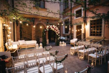 An intimate wedding venue between brick buildings. There are twinkle lights and vines strung above an altar with white and red flowers.